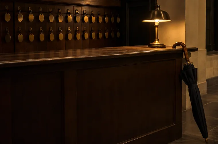 A gallery coat-check counter at night: brass number tags on the wall, a lamp on the desk and an umbrella leaning by it