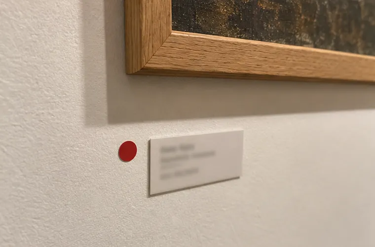 A red dot sticker beside a small museum label under an oak frame on a white wall